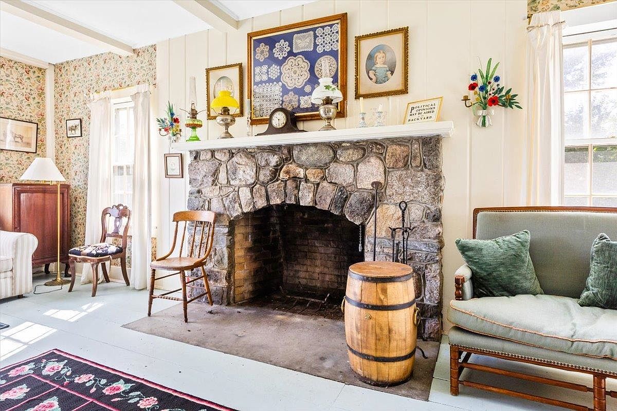 This cozy living room features a prominent stone-clad fireplace as its central focal point, topped with a wooden mantel displaying various vintage decor and framed artwork. The room is furnished with a light-toned sofa, a wooden barrel side table, and classic wooden chairs, all set against a backdrop of patterned wallpaper and light-colored walls. The perspective is a straight-on shot that captures the rustic, traditional charm of the space, highlighting the hearth and the surrounding seating area.
