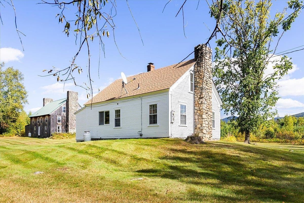This side view captures a white-sided farmhouse set on a gentle, grassy slope under a clear blue sky. A prominent stone-clad chimney rises along the side of the home, adding a rustic architectural element, while a second, older wooden structure stands in the background. The scene is framed by tree branches in the foreground, emphasizing the rural, peaceful setting of the property.