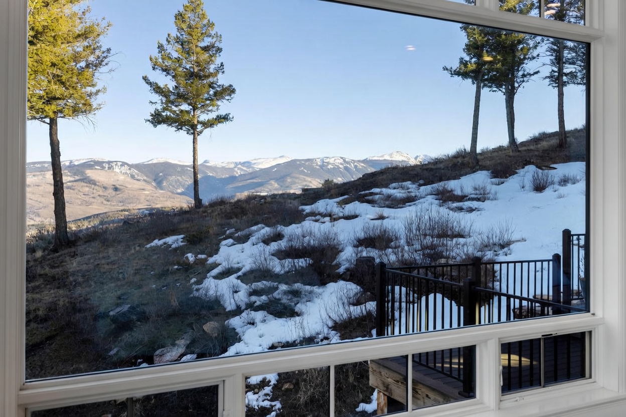 This interior perspective looks out through a large window frame, showcasing a breathtaking panoramic view of snow-dusted mountains and a rugged, natural landscape. In the foreground, a dark metal railing of a deck is visible, bridging the indoor space with the expansive outdoor scenery. The composition emphasizes the connection between the home's interior and the serene, mountainous environment, highlighting the property's scenic location.