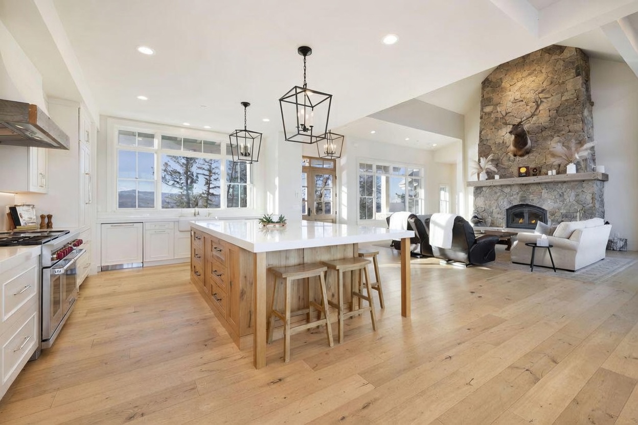 This open-concept space features a large kitchen island with a light-toned stone countertop and wood cabinetry, serving as the centerpiece of the room. Beyond the island, a living area is anchored by a prominent stone-clad chimney breast and a mounted animal trophy, creating a rustic yet refined atmosphere. The room is bathed in natural light from large windows and is finished with light wood flooring, offering a seamless flow between the cooking and relaxation zones.