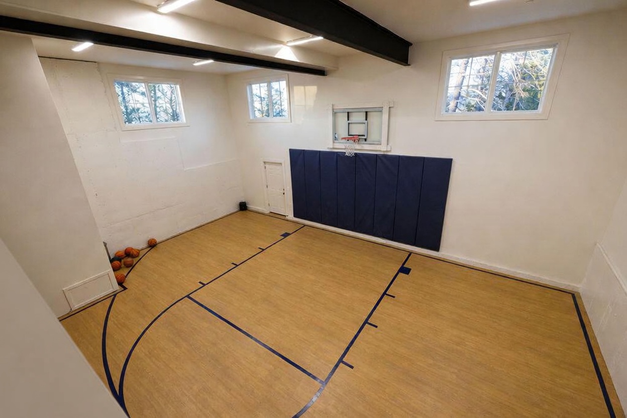 This indoor home gym features a dedicated basketball court area with light wood-look flooring and painted court markings. A wall-mounted basketball hoop is centered above a dark blue padded wall protector, while several basketballs are neatly stored in the corner. The space is illuminated by overhead lighting and includes multiple windows that provide natural light, creating an active and functional recreational area.