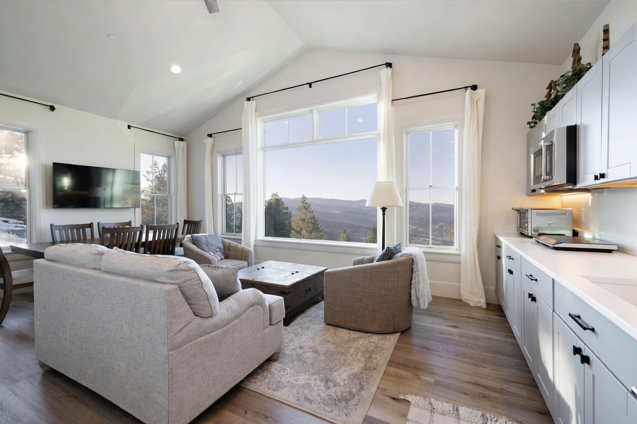 This open-concept living area features a light-toned sofa and two armchairs arranged around a rustic wooden coffee table on a patterned area rug. Large windows frame a panoramic view of the surrounding mountains, filling the space with natural light. To the right, a clean-lined kitchen with light-colored cabinetry and modern appliances seamlessly integrates into the living space, creating an airy and inviting atmosphere.