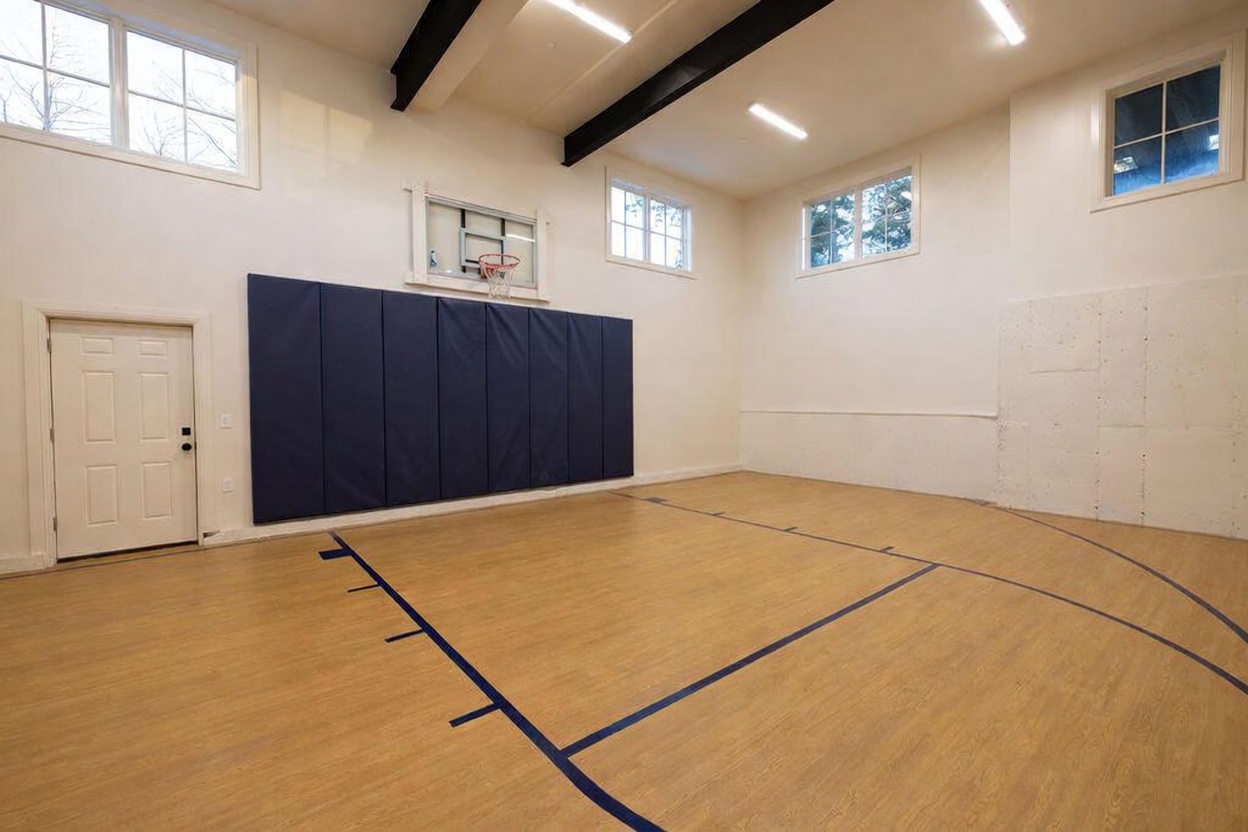 This indoor sports court features light wood flooring with painted blue court markings and a wall-mounted basketball hoop protected by dark blue padded wall mats. The space is illuminated by multiple overhead light fixtures and features several windows that provide natural light, creating an open and airy atmosphere. The room is designed for recreational activity, with a clean, functional aesthetic and high ceilings accented by dark structural beams.