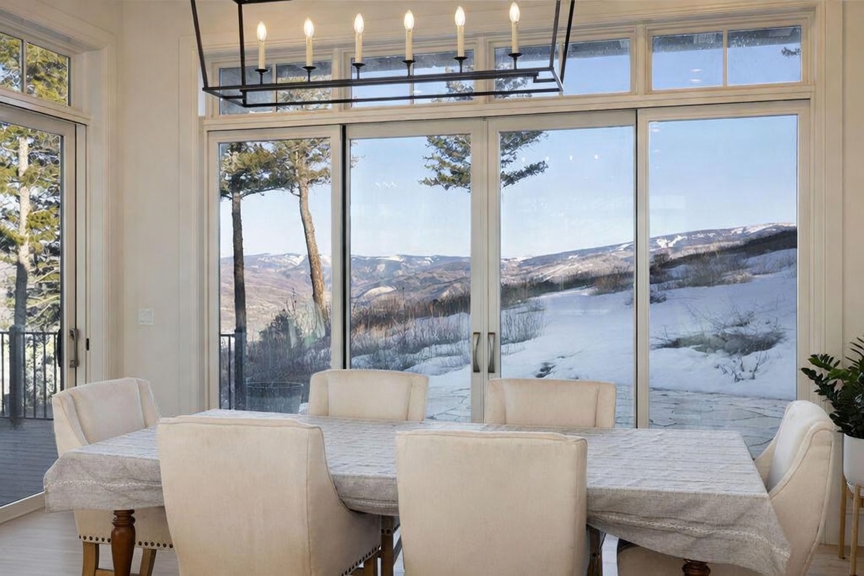This bright dining room features a large table draped in a neutral-toned cloth, surrounded by upholstered chairs. A prominent rectangular chandelier with six exposed-bulb lights hangs above the table, drawing the eye toward the expansive wall of glass. The room offers a stunning, unobstructed view of a snow-covered mountain landscape through large windows that span the width of the wall.