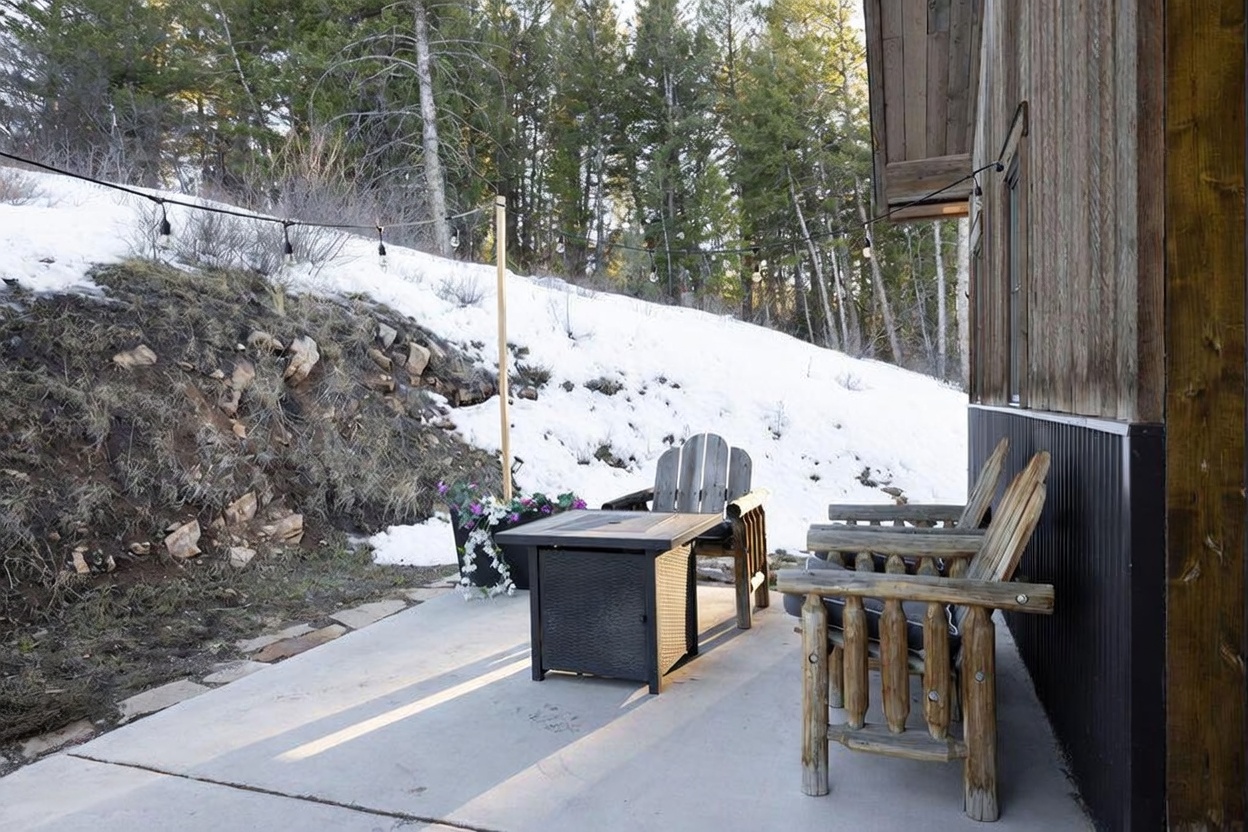 This outdoor patio space features a concrete surface furnished with a square fire pit table and rustic log-style wooden chairs. String lights are draped above the seating area, which is set against a backdrop of a snow-covered hillside and a dense forest. The side of the home, finished in vertical wood siding and dark metal paneling, frames the right side of the scene.