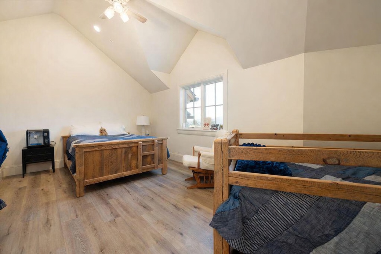 This bedroom features a vaulted ceiling and light wood-look flooring, creating an airy and spacious atmosphere. The room is furnished with a wooden bed frame and a bunk bed, complemented by a rocking chair positioned near a window that allows for natural light. The neutral wall color and rustic wood furniture provide a cozy, inviting aesthetic suitable for a guest or children's room.