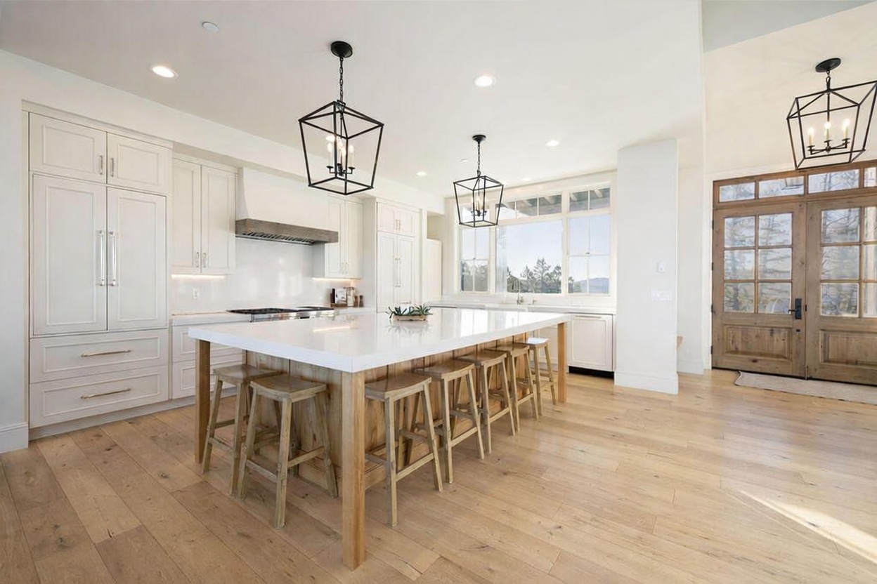This bright, modern kitchen features a large central island with seating for four, topped with a white surface and supported by a light wood base. The space is equipped with white cabinetry, a professional-style range, and three prominent black geometric pendant lights hanging above the island. Large windows provide natural light and a view of the outdoors, while a set of wooden double doors leads to an entryway, creating an open and airy atmosphere.