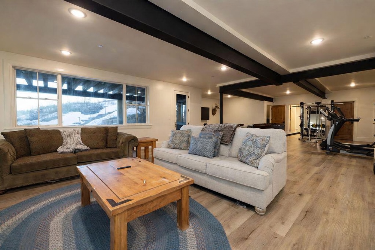 This spacious, open-concept living area features light wood-look flooring and a prominent dark structural beam overhead. The room is furnished with a large, light-colored sofa and a brown sofa, centered around a rustic wooden coffee table on a circular blue rug. Large windows provide ample natural light and a view of a snowy outdoor landscape, while a home gym area is visible in the background.