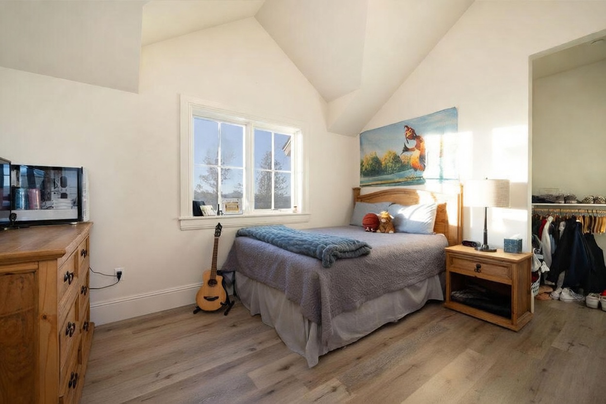 This cozy bedroom features a vaulted ceiling and light wood-look flooring, creating an airy and inviting atmosphere. A queen-sized bed with a blue quilt is centered against a wall adorned with a large, colorful character poster, while a wooden dresser and matching nightstand provide ample storage. A window allows natural light to fill the space, and an open closet area with hanging clothes is visible to the right.
