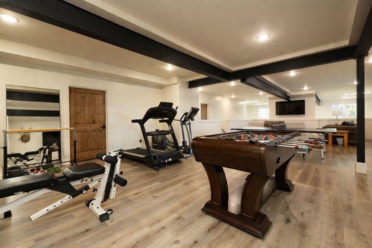 This spacious, multi-functional basement serves as both a home gym and a recreation area, featuring light wood-look flooring and exposed dark ceiling beams. The room is equipped with a treadmill, an elliptical machine, a weight bench, and a foosball table, with a ping-pong table visible in the background. The open-concept layout creates an inviting space for fitness and leisure, illuminated by recessed lighting throughout.