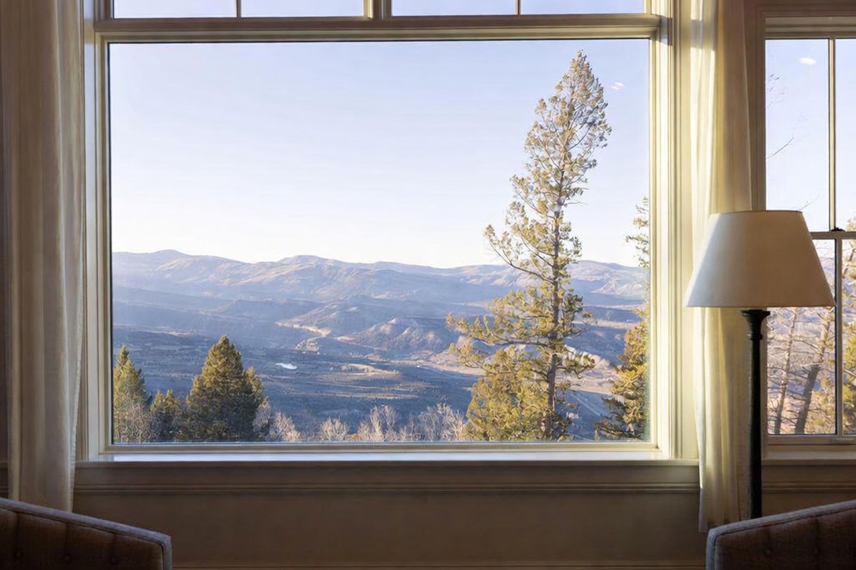 This interior view highlights a large window framing a breathtaking, expansive mountain vista. The scene includes a portion of a lamp with a neutral shade on the right and the tops of two upholstered chairs in the foreground, suggesting a comfortable seating area. The composition emphasizes the connection between the indoor living space and the natural beauty of the surrounding landscape.