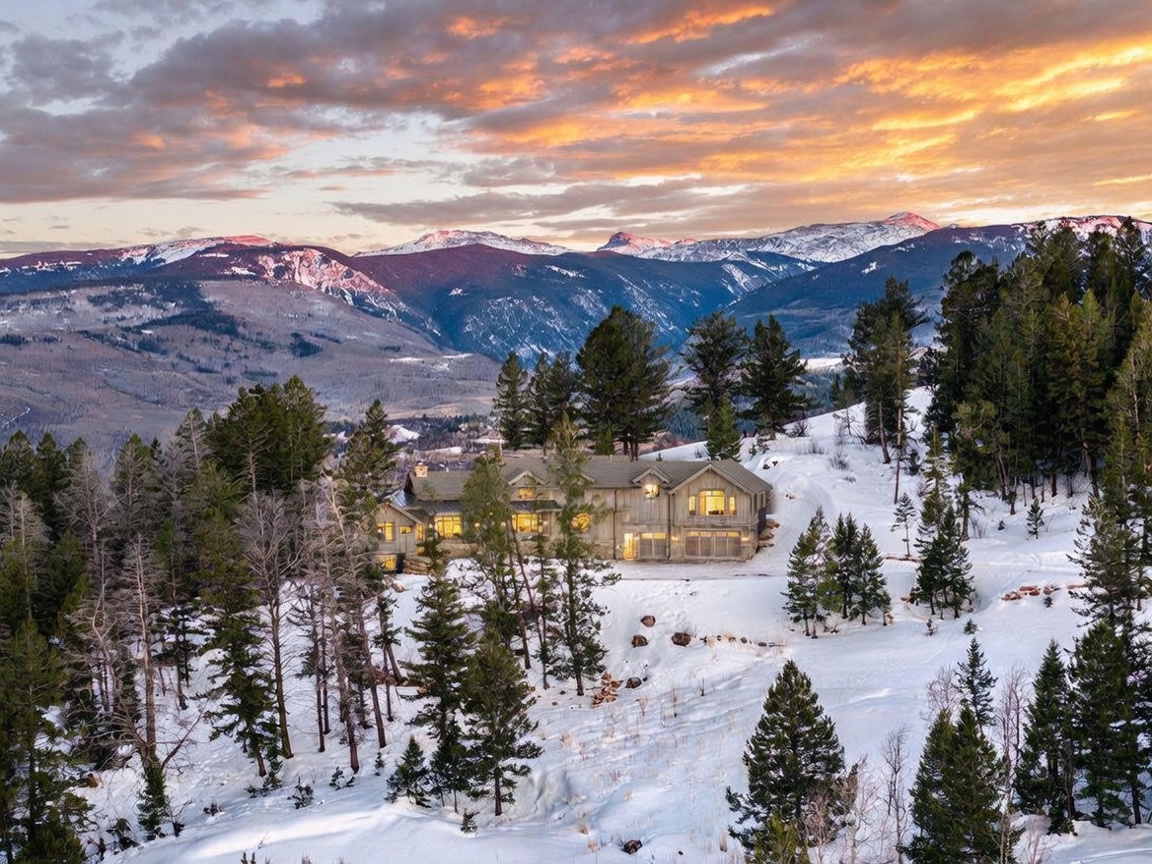 This aerial perspective captures a sprawling mountain estate nestled within a snow-covered, forested landscape at sunset. The home features a rustic, multi-gabled architectural style with warm interior lighting glowing through numerous windows, contrasting beautifully against the cool tones of the winter twilight. In the background, majestic, snow-capped mountain peaks rise against a vibrant, cloud-streaked sky, creating a serene and picturesque setting.
