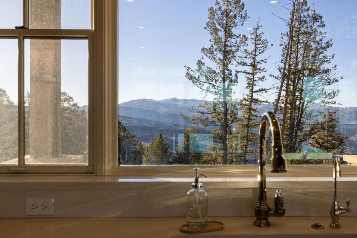 This interior view captures a kitchen sink area positioned directly in front of a large window that frames a scenic mountain landscape. The scene features a polished metal faucet, a glass soap dispenser, and a clean, light-toned countertop surface. The perspective emphasizes the connection between the indoor space and the expansive natural views outside.