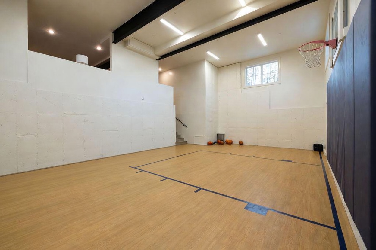 This indoor sports court features light-toned wood flooring with painted blue court markings and a basketball hoop mounted on the wall. The space is enclosed by white walls, with a section of dark padded wall protection near the hoop, and includes a small set of stairs leading to an upper level. The room is illuminated by recessed lighting and a window, creating a functional and recreational atmosphere.