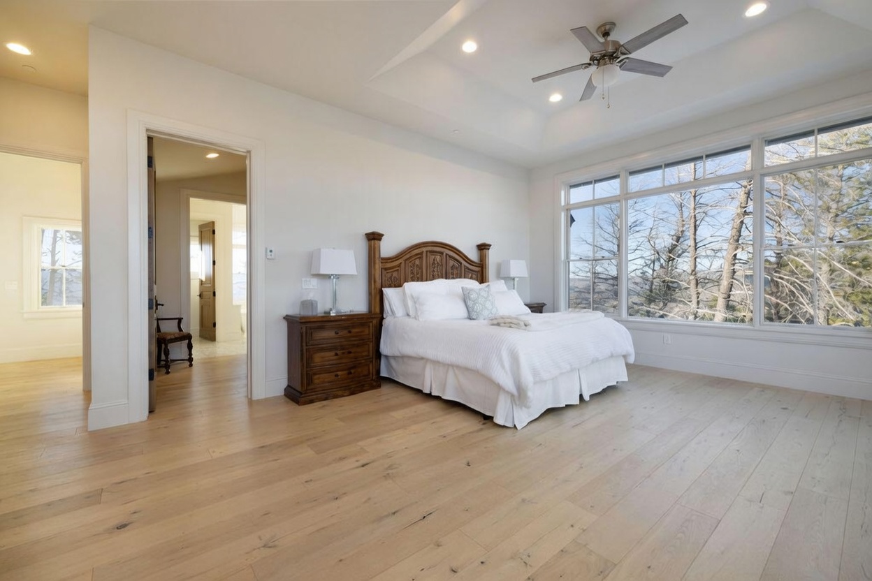This spacious primary bedroom features light wood flooring and a large, ornate wooden headboard as the focal point. A wall of expansive windows provides abundant natural light and a scenic view of the surrounding trees, while a tray ceiling with a central fan adds architectural interest. The room connects to an adjacent hallway, creating an open and airy atmosphere.