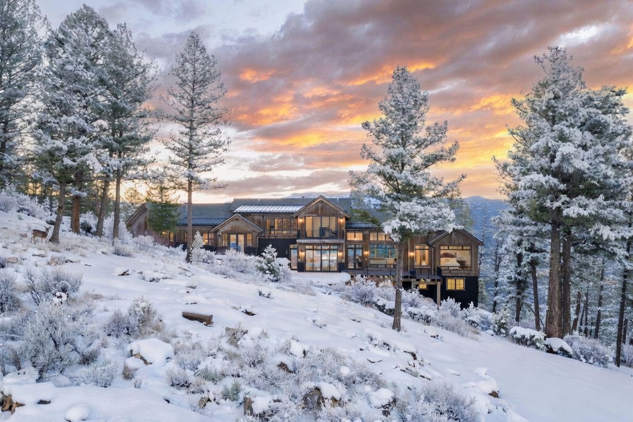 This stunning mountain estate is captured at dusk, showcasing a multi-level architectural design with a blend of dark wood siding and expansive glass windows. The home is nestled into a snow-covered hillside surrounded by frost-dusted pine trees, creating a serene and luxurious winter atmosphere. The warm interior lighting glows through the large windows, contrasting beautifully with the vibrant orange and purple hues of the sunset sky.