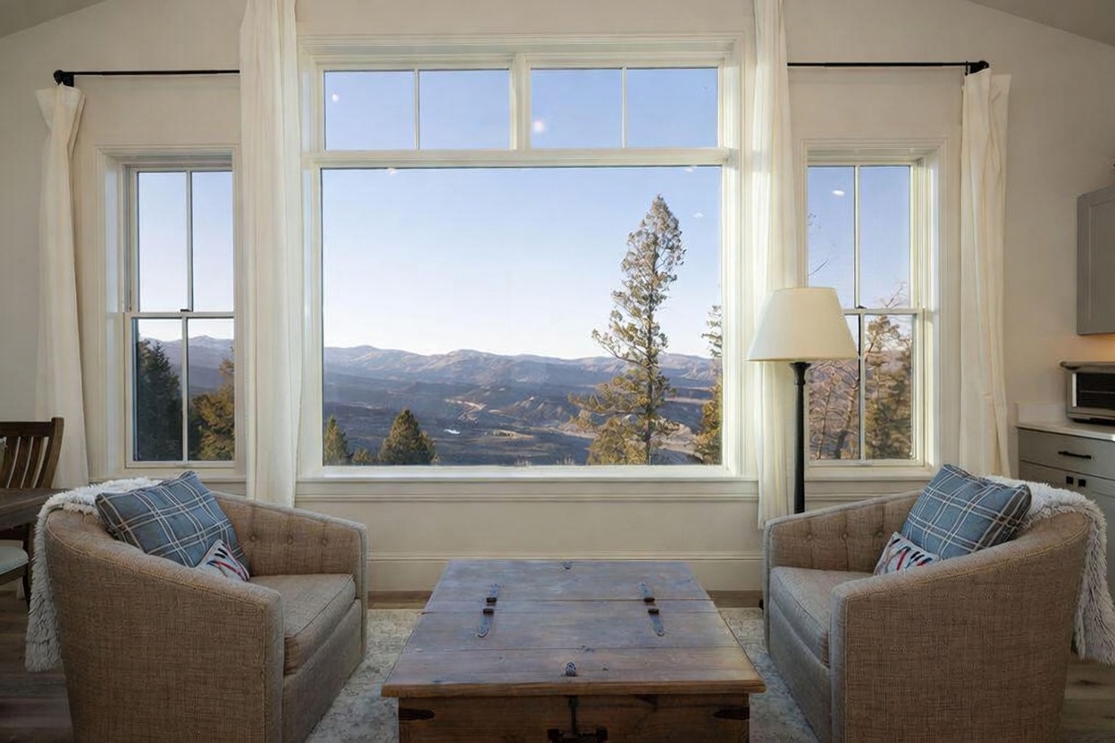 This cozy living area features two plush armchairs with plaid accent pillows facing a rustic wooden coffee table. A large window serves as the focal point, framing a breathtaking panoramic view of distant mountain ranges and evergreen trees. The space is framed by soft white curtains and includes a floor lamp, creating a serene and inviting atmosphere perfect for taking in the scenery.