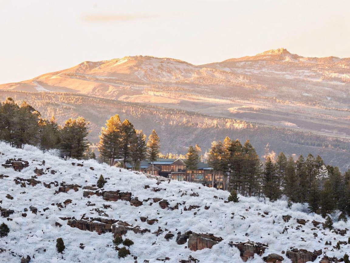 This wide-angle shot captures a luxurious mountain residence nestled into a snowy, rugged hillside. The home features a multi-level design with extensive glass, blending seamlessly into the surrounding pine forest and rocky terrain. The golden hour light illuminates the peaks in the background, creating a dramatic and serene atmosphere for this secluded estate.