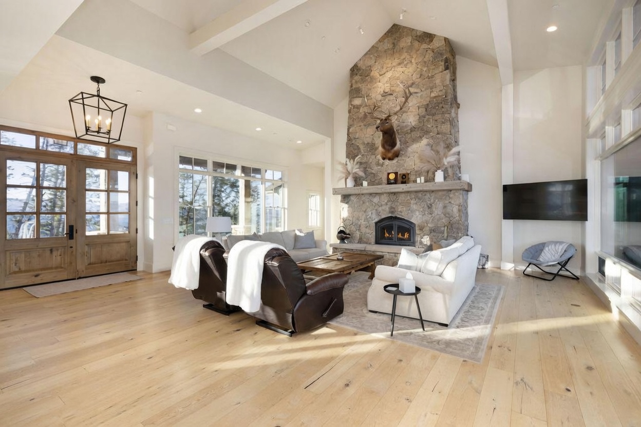 This spacious living room features a vaulted ceiling with exposed beams and light wood flooring throughout. A prominent stone-clad chimney breast serves as the focal point, adorned with a mounted elk head above the mantel and a fireplace below. The room is furnished with a pair of leather recliners and a white sofa, all arranged around a rustic wood coffee table, while large windows provide ample natural light and views of the outdoors.