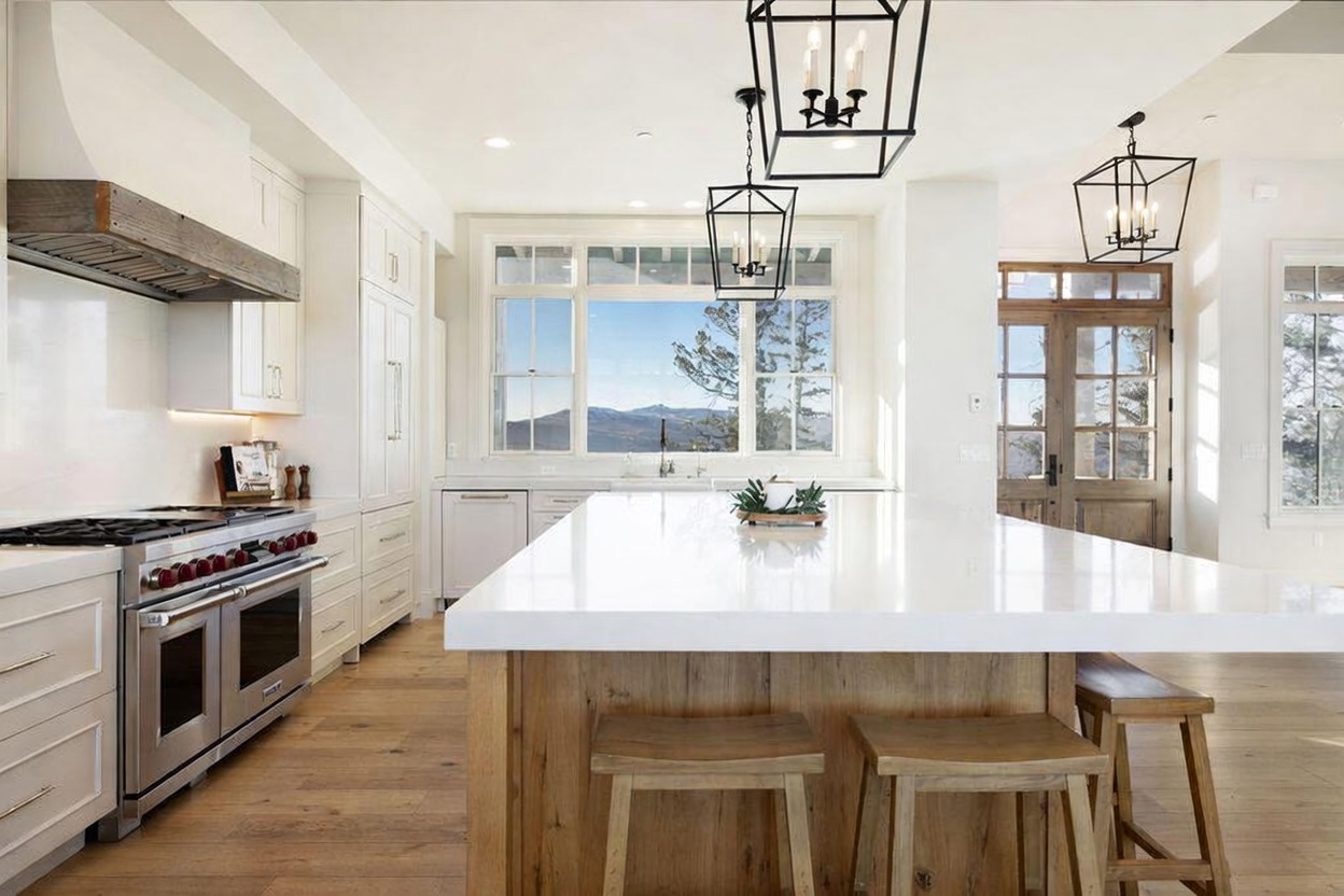 This bright, modern kitchen features a large central island with a light-toned wood base and a white countertop, complemented by three wooden stools. White cabinetry and a professional-grade stainless steel range are set against a clean, minimalist backdrop, while large windows above the sink provide a scenic view of the mountains. The space is illuminated by three black geometric pendant lights, creating a sophisticated and airy atmosphere.