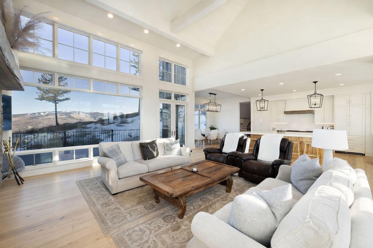 This expansive, open-concept living area features a vaulted ceiling and a wall of large windows that frame a scenic, snow-covered mountain landscape. The space is furnished with two light-colored sofas and a pair of dark leather recliners arranged around a rustic, weathered wood coffee table on a patterned area rug. Beyond the living area, the space transitions into a bright kitchen with a large island and pendant lighting, creating a cohesive and airy atmosphere perfect for entertaining.