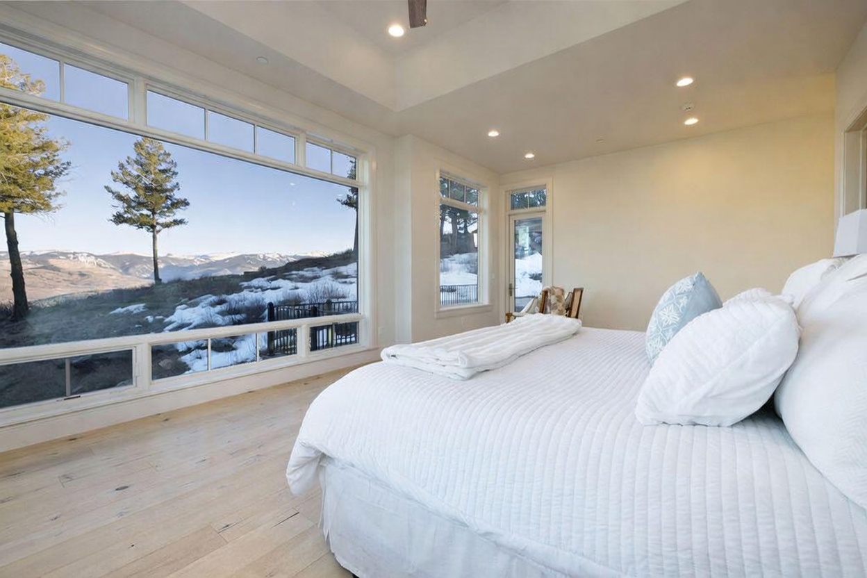 This bright and airy primary bedroom features light wood flooring and a large, plush bed dressed in crisp white linens. A massive expanse of glass provides an unobstructed, panoramic view of a snow-dusted mountain landscape, seamlessly blending the interior with the natural surroundings. The room is finished with recessed ceiling lighting and a neutral color palette, creating a serene and luxurious atmosphere.