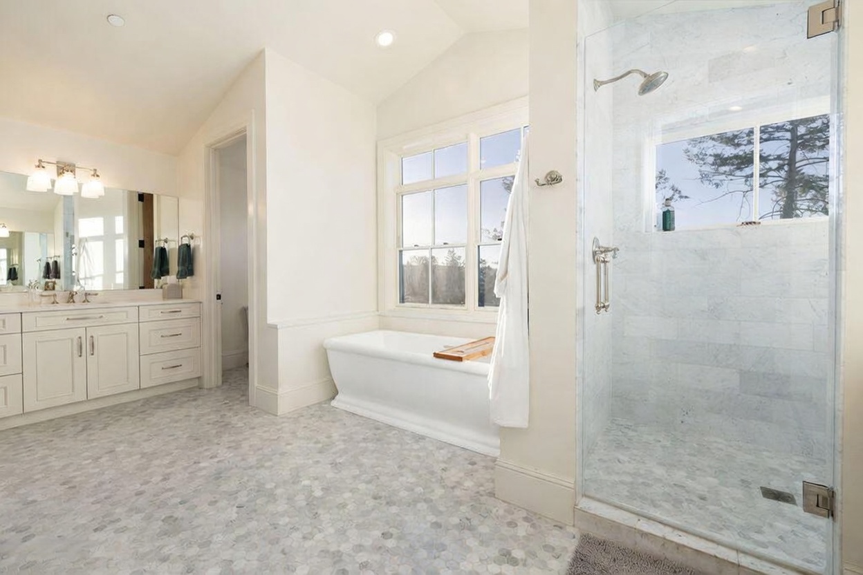 This bright primary bathroom features a freestanding soaking tub positioned beneath a large window, flanked by a glass-enclosed walk-in shower with light-toned tile walls. A double vanity with white cabinetry and a wide mirror provides ample storage and counter space, while the floor is finished in a decorative hexagonal patterned tile. The room is bathed in natural light, creating a clean, airy, and spa-like atmosphere.