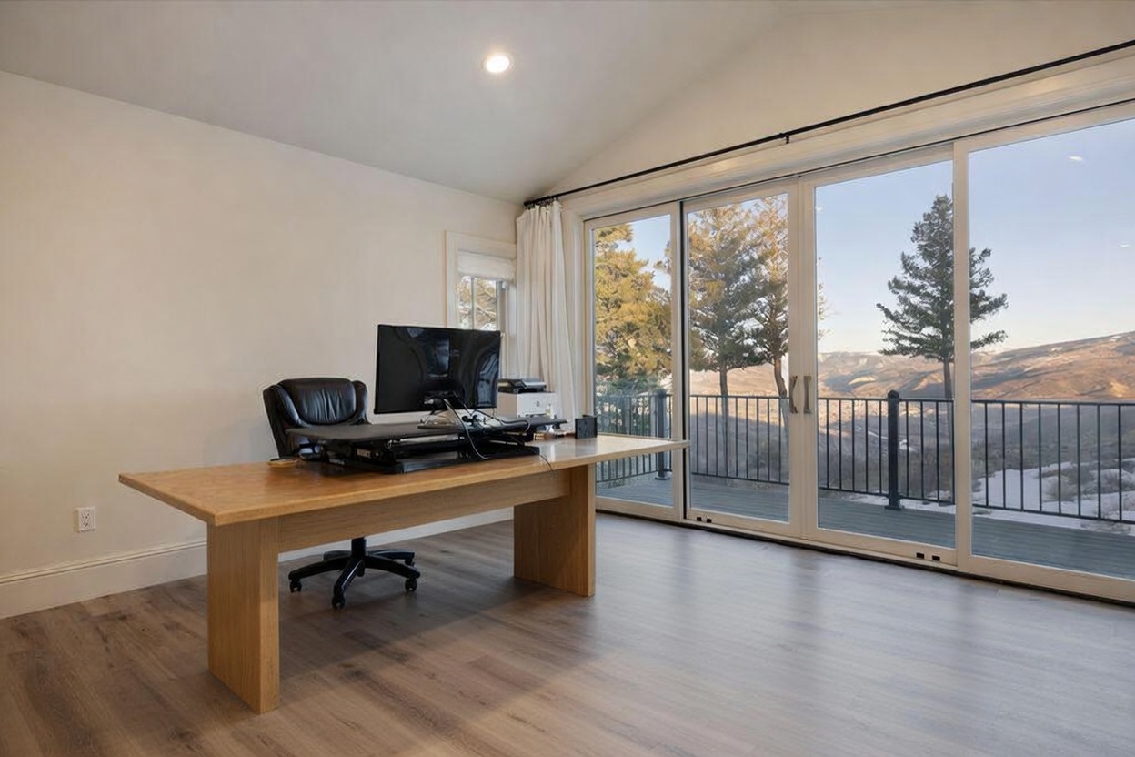 This bright home office features a large light wood desk positioned to face a wall of expansive glass sliding doors that provide panoramic mountain views. The room is anchored by a plush, light-colored area rug and includes a black ergonomic office chair and a computer setup. The vaulted ceiling and neutral wall tones create an airy, professional atmosphere perfect for focused work.