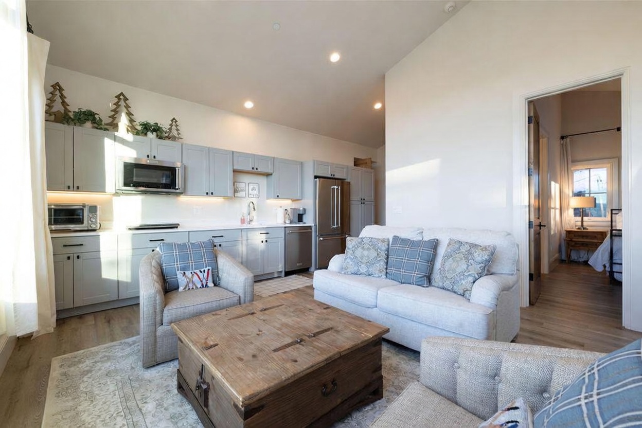 This open-concept living area features a cozy seating arrangement with a light-colored sofa and two armchairs centered around a rustic wooden coffee table. The space seamlessly integrates with a kitchen area, which is equipped with light blue cabinetry, stainless steel appliances, and a clean, neutral aesthetic. The room is bright and airy, with light wood flooring and a view leading into an adjacent bedroom.