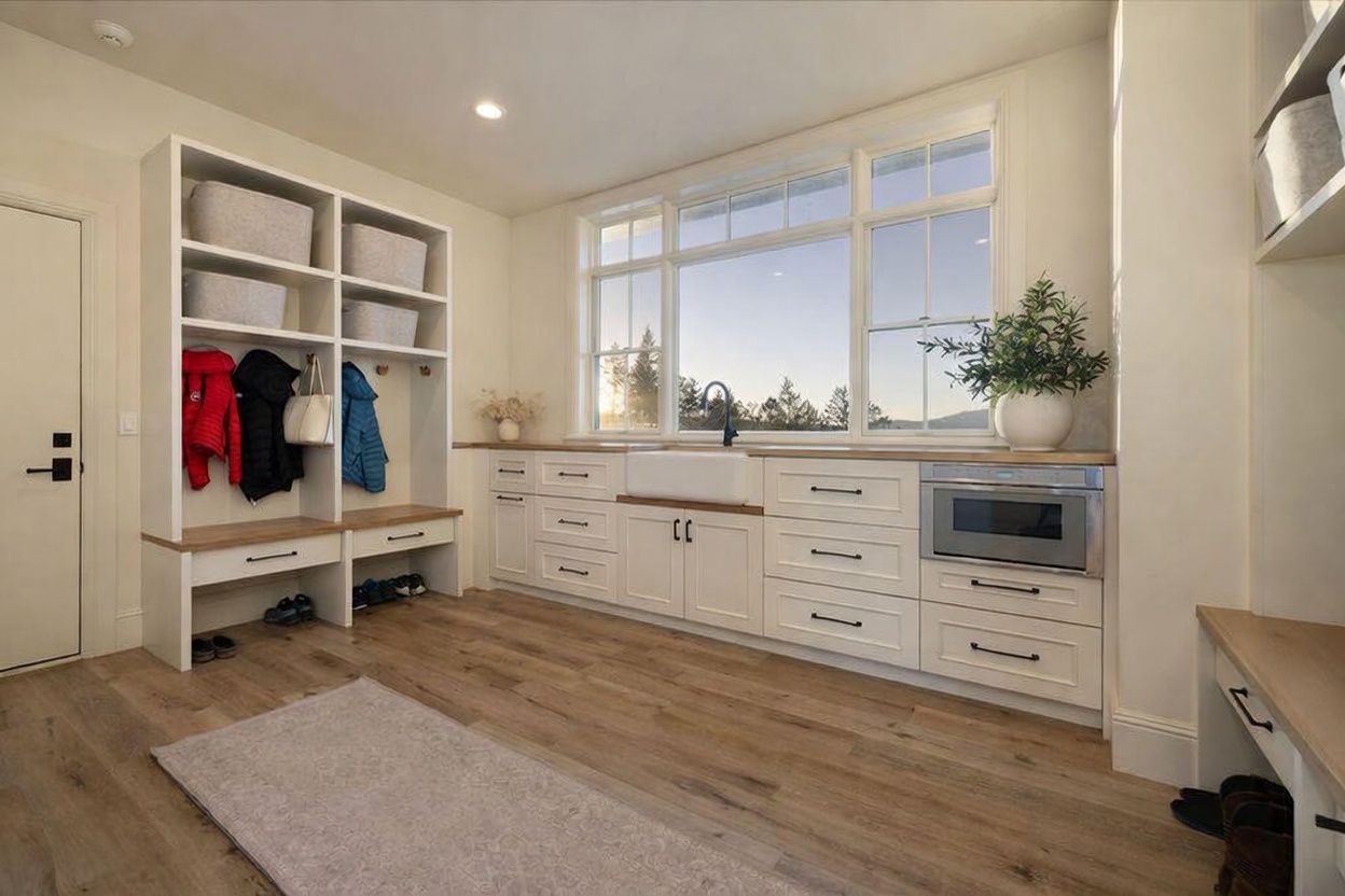 This bright and functional mudroom or laundry space features custom white built-in cabinetry with integrated bench seating and open storage cubbies. A farmhouse-style sink is set into a light wood countertop, complemented by a built-in microwave and large windows that provide ample natural light and a view of the outdoors. The room is finished with light wood-tone flooring and a neutral area rug, creating a clean and organized aesthetic.