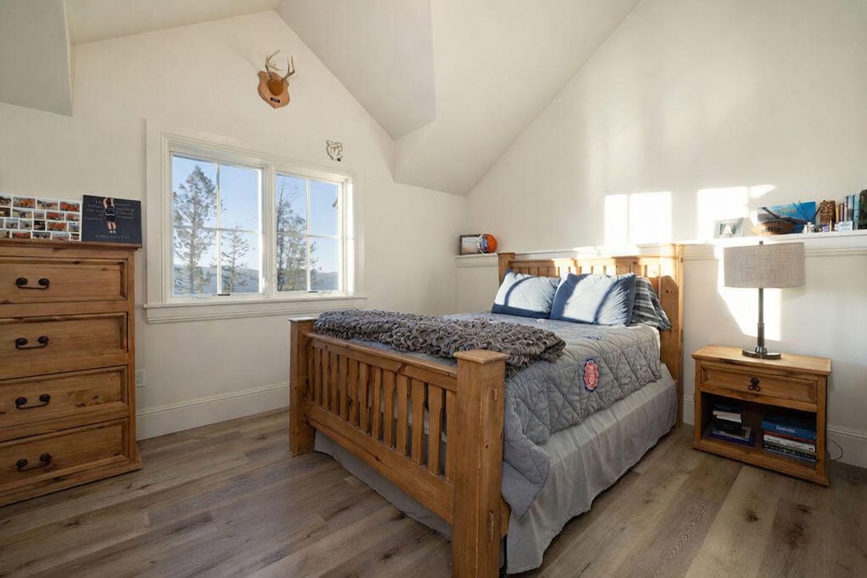 This cozy bedroom features a wooden bed frame with a quilted gray bedding set and a matching wooden dresser and nightstand. The room is illuminated by natural light streaming through a window that offers a scenic view of the surrounding trees and hills. A vaulted ceiling adds a sense of height and openness to the space, which is decorated with personal touches like a wall-mounted deer mount and a shelf above the bed.