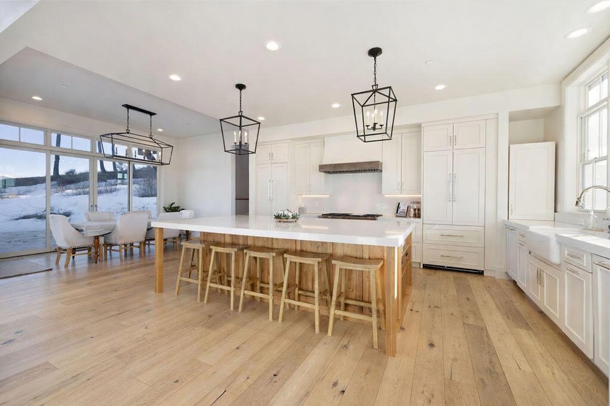 This bright, open-concept kitchen features a large central island with a light-toned wood base and a white countertop, complemented by four wooden bar stools. The space is anchored by white cabinetry, a custom range hood, and light wood flooring that extends into the adjacent dining area. Large glass doors provide an expansive view of a snowy outdoor landscape, filling the room with natural light.