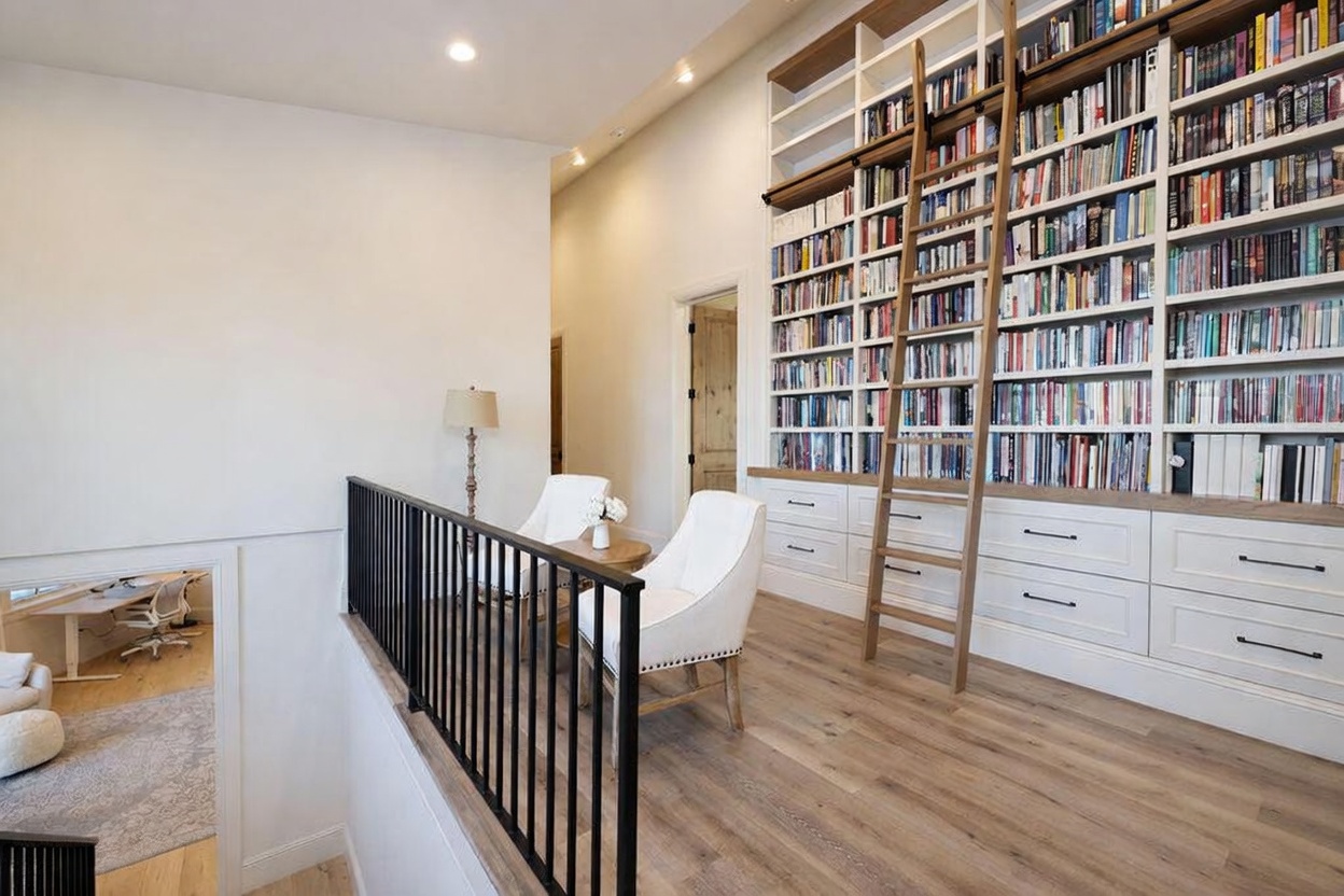 This elevated landing area features a striking library wall with built-in shelving and a rolling ladder, creating a sophisticated reading nook. Two white armchairs are positioned near a small side table, overlooking a lower level visible through a black metal railing. The space is finished with light wood flooring and neutral walls, offering a bright and airy atmosphere.