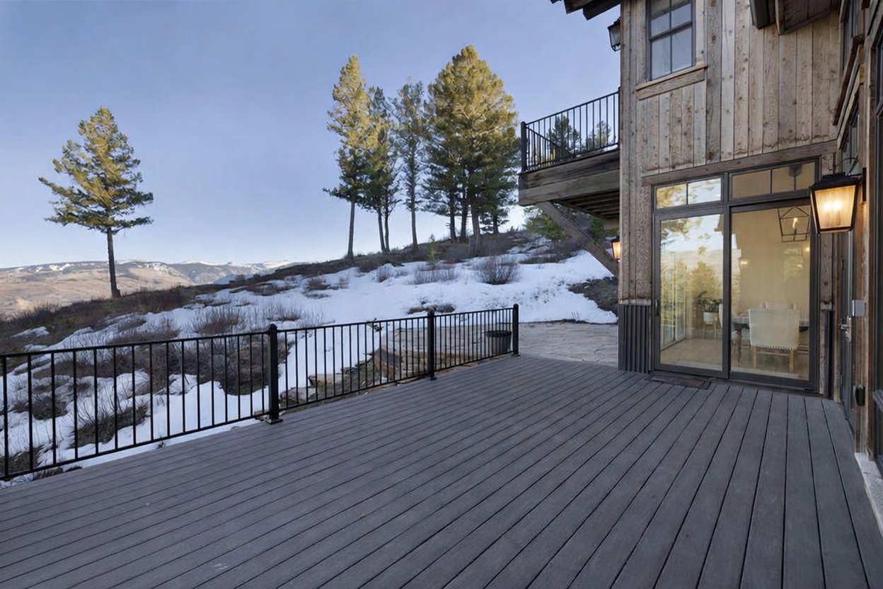 This image captures a spacious wooden deck featuring a black metal railing that overlooks a snowy, mountainous landscape. The deck connects directly to the home's interior through large glass sliding doors, which reveal a glimpse of a dining area inside. The rustic wood siding of the house and the expansive, scenic view create a serene and inviting atmosphere.