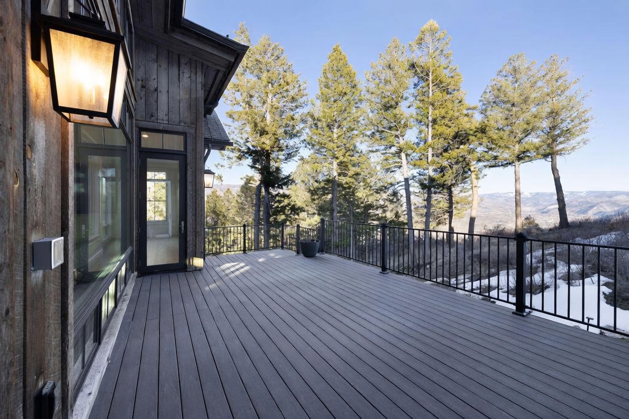 This spacious, elevated deck features dark-toned composite decking and a sleek black metal railing that provides an unobstructed view of the surrounding mountain landscape. The exterior of the home is clad in vertical wood siding, complemented by a large glass door and a modern wall-mounted light fixture. The perspective captures the expansive outdoor living space, emphasizing the connection between the home's rustic aesthetic and the natural, scenic environment.