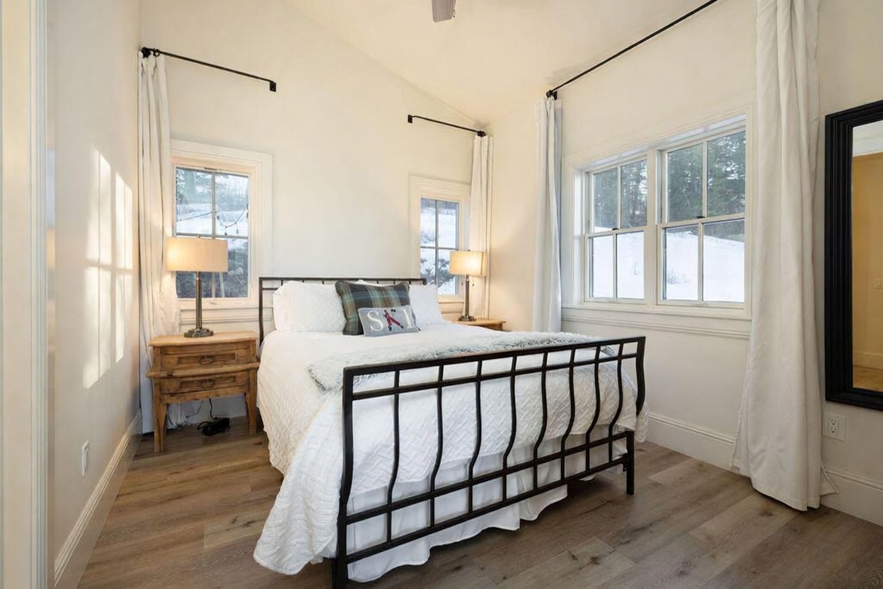 This cozy bedroom features a black metal-framed bed dressed in white linens and a plaid accent pillow, set against a backdrop of light-toned walls and wood-look flooring. Two windows with white curtains allow natural light to fill the space, revealing a snowy landscape outside. The room is furnished with a rustic wooden nightstand and a matching lamp, creating a warm and inviting atmosphere.