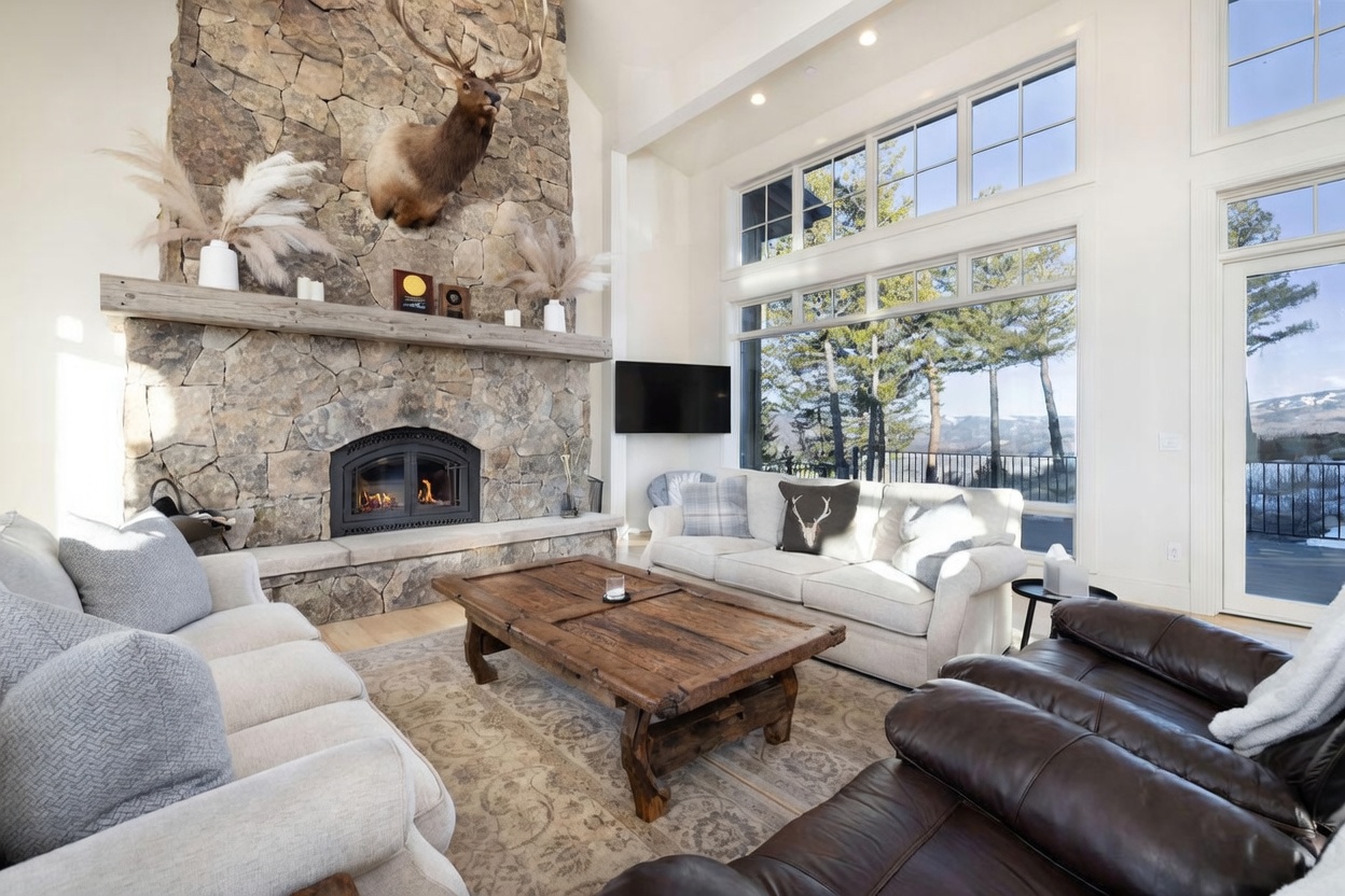 This spacious living room features a prominent stone-clad chimney breast with a mounted elk head above the mantel, creating a rustic mountain aesthetic. The room is anchored by a large, weathered wood coffee table and comfortable seating, while expansive windows provide abundant natural light and scenic views of the surrounding landscape. The high ceilings and open layout contribute to an airy, inviting atmosphere perfect for relaxation.