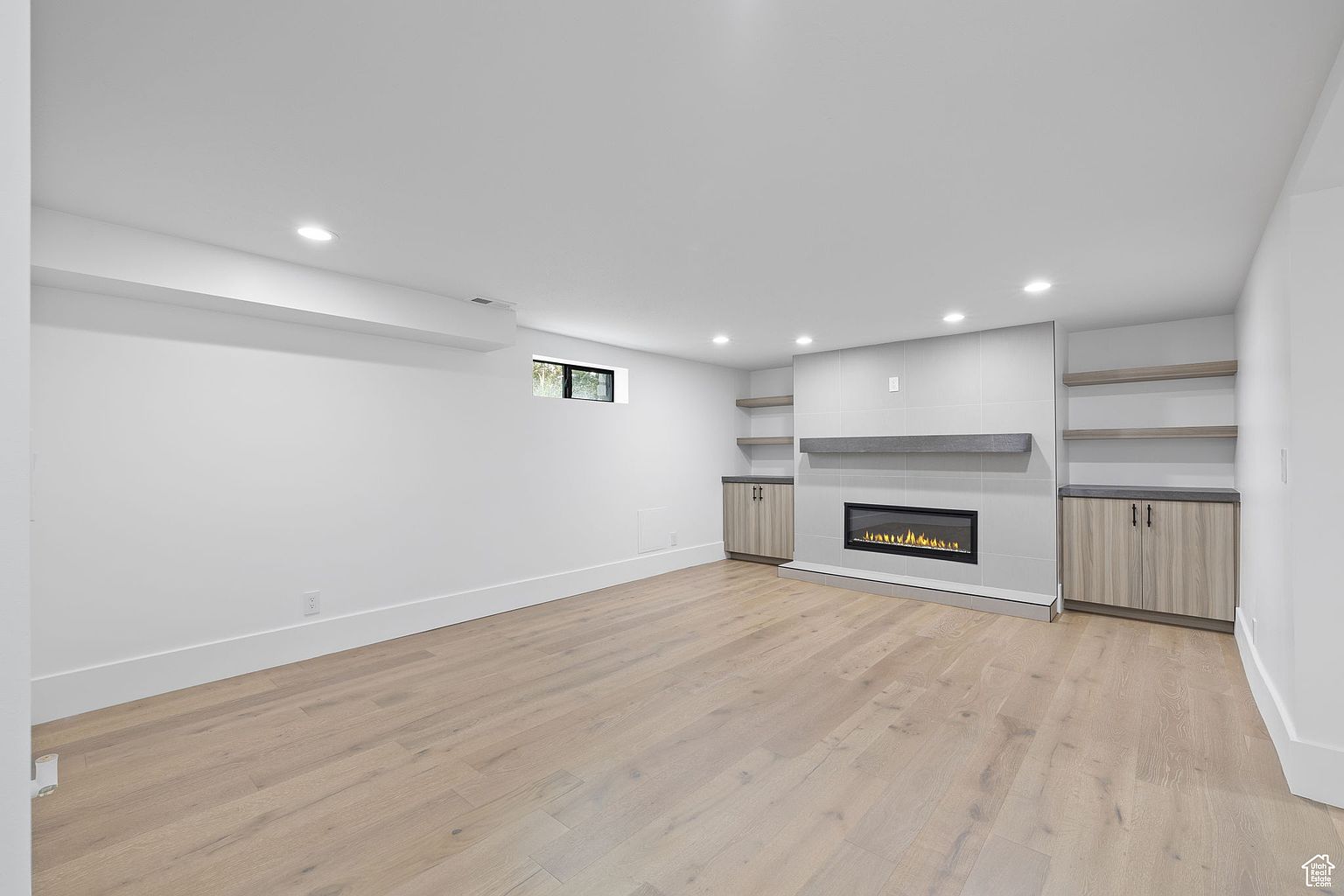 This is an interior shot of a modern living room featuring light wood flooring and white walls. A sleek, linear fireplace is the focal point, flanked by built-in cabinets and shelving. The room is well-lit with recessed lighting, creating a bright and inviting atmosphere.