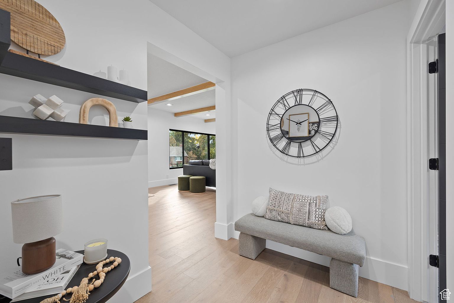 This interior shot showcases a bright and modern hallway with light wood flooring and white walls. A decorative clock hangs above a gray upholstered bench, adding a touch of elegance. The hallway leads into a living area with exposed wooden beams, creating a seamless transition between spaces.