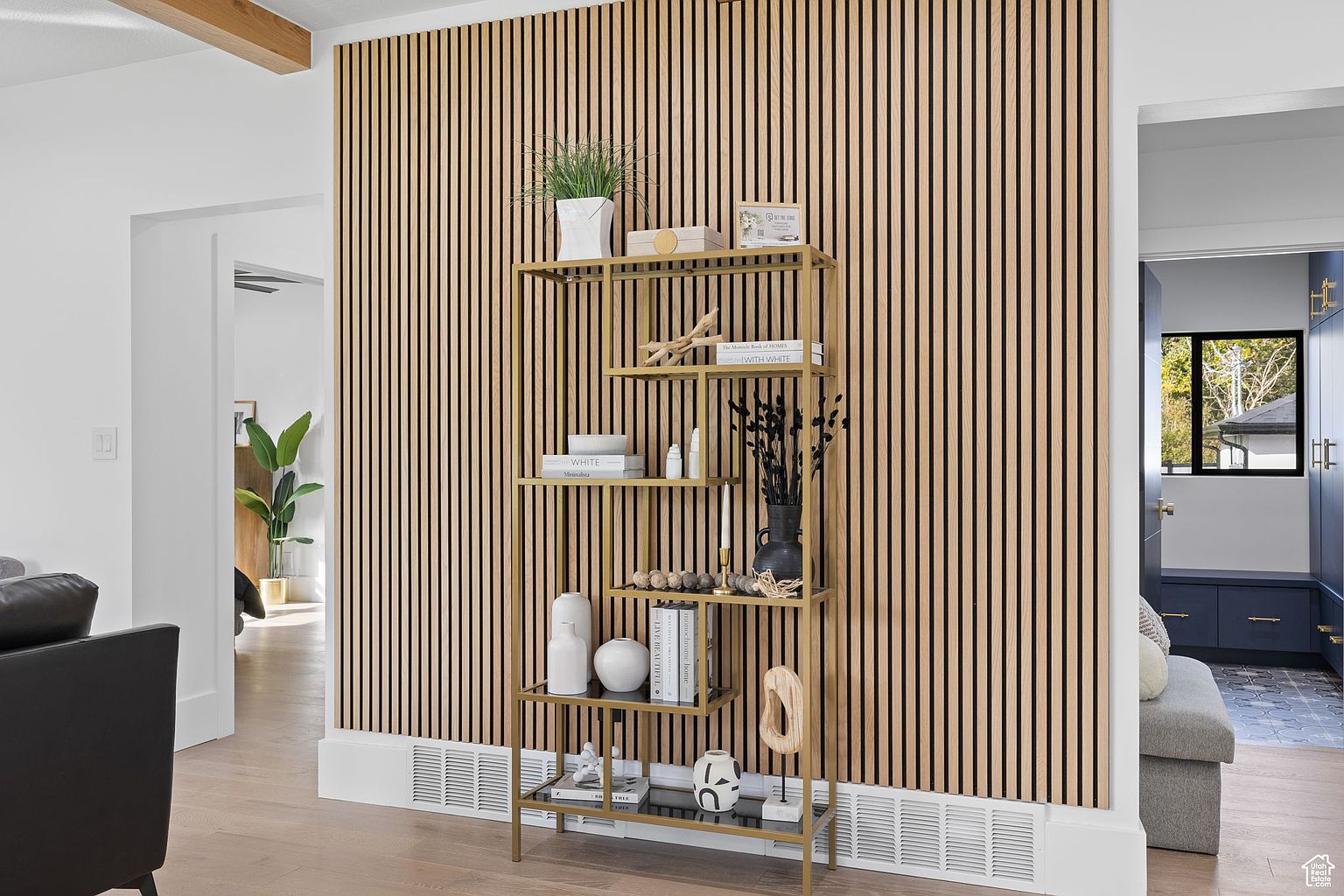 This interior shot showcases a stylish living room with a focus on modern design. A gold-toned shelving unit displays decorative items against a backdrop of vertically paneled walls, adding texture and visual interest. The room features hardwood flooring and glimpses into adjacent spaces, creating an open and inviting atmosphere.