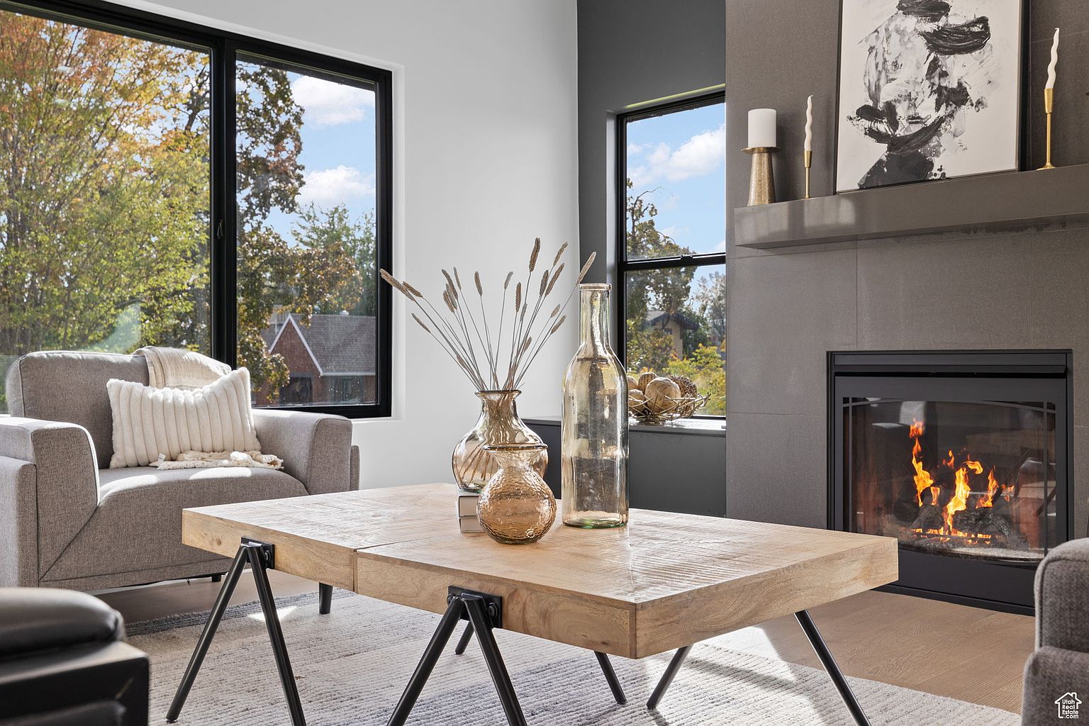 This is a cozy living room featuring a modern fireplace with a gray tile surround and a black firebox. A light wood coffee table with black metal legs sits on a patterned rug, surrounded by a gray armchair and sofa. Large windows provide natural light and views of the outdoors, creating a warm and inviting atmosphere.