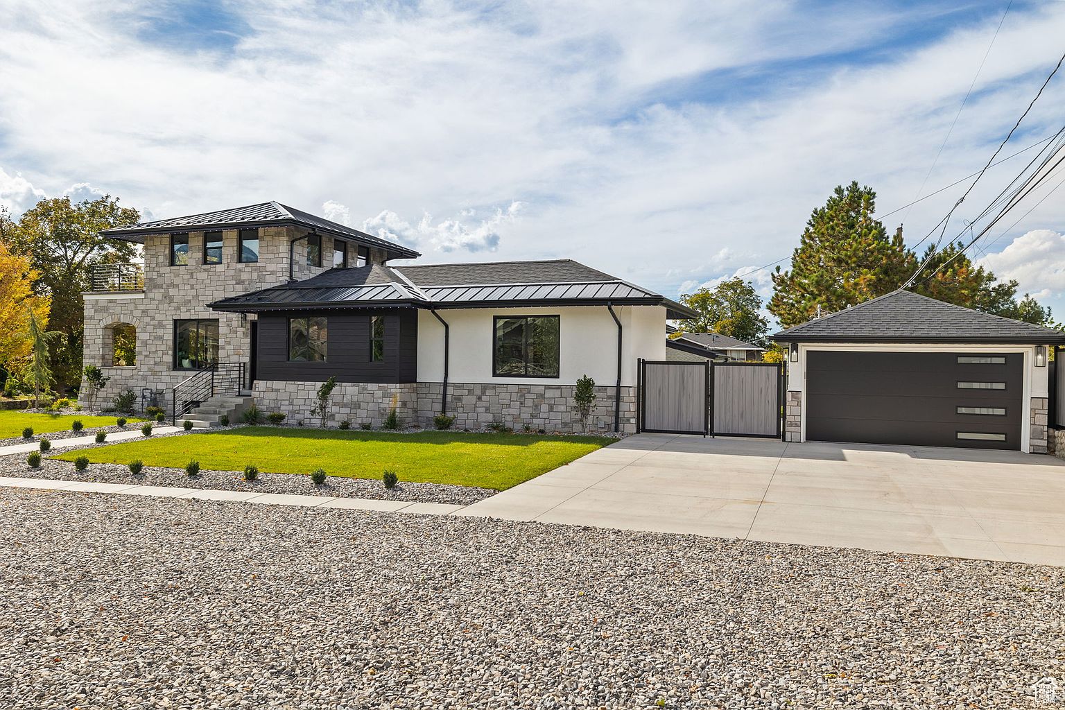 This is a front exterior view of a modern home with a combination of stone and dark wood siding. The house features a well-manicured lawn, a gravel driveway, and a detached garage with a matching design. The overall impression is one of sophisticated curb appeal and contemporary design.