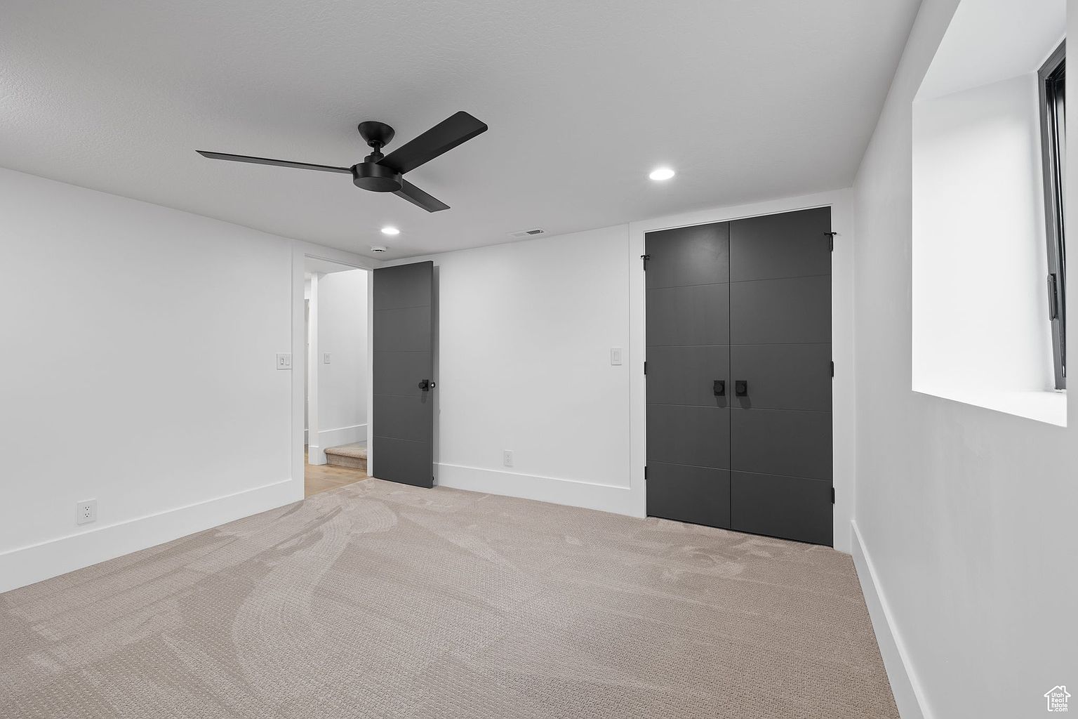 This is a well-lit bedroom featuring neutral-toned carpet and white walls, creating a bright and airy atmosphere. The room includes a black ceiling fan and two dark gray doors, one single and one double, adding a modern contrast. A window provides natural light, enhancing the room's appeal for potential buyers.