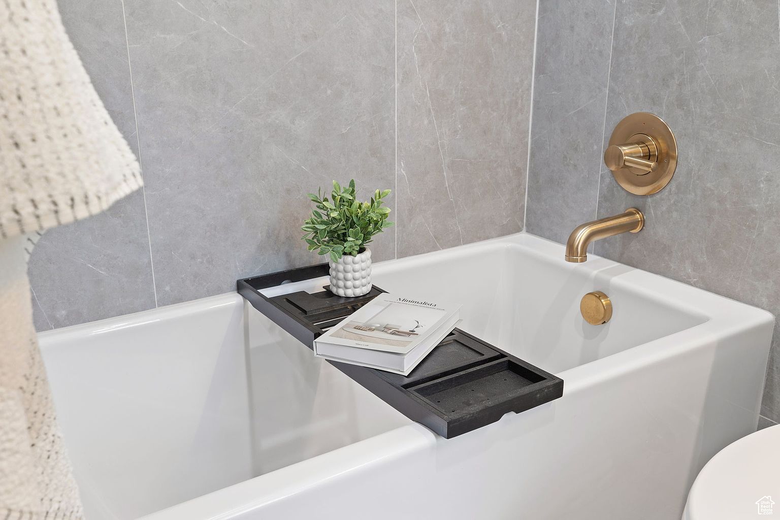 This is a close-up shot of a primary bathroom featuring a modern, white bathtub with gold fixtures. A black bath tray holds a small potted plant and a book, adding a touch of elegance and relaxation. The gray tiled walls provide a clean and sophisticated backdrop.