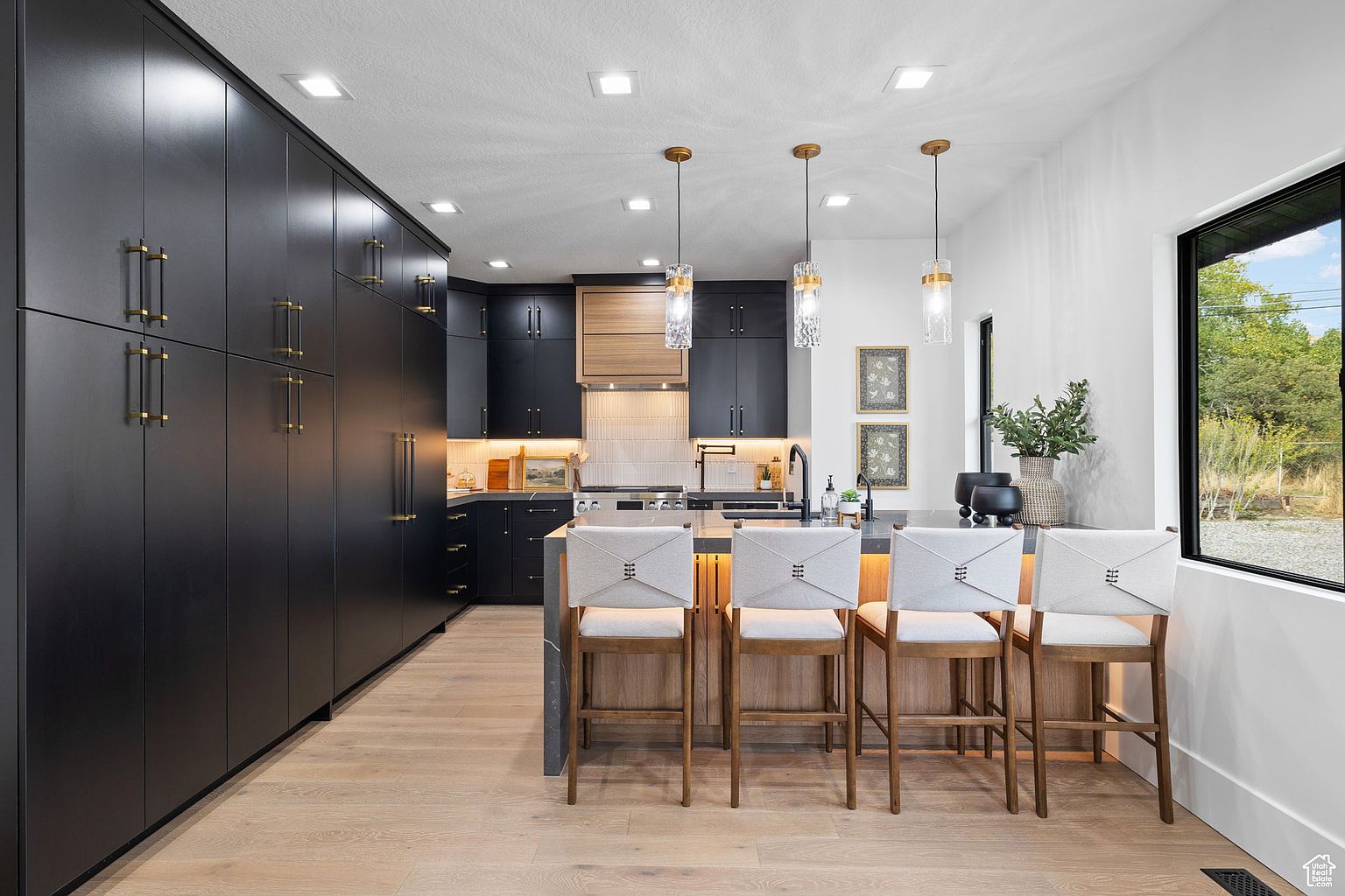 This is a modern kitchen featuring sleek, dark cabinetry with gold hardware, complemented by light wood flooring. A central island with bar seating is illuminated by pendant lights, and a large window provides natural light and a view of the outdoors. The kitchen exudes a sophisticated and contemporary aesthetic.