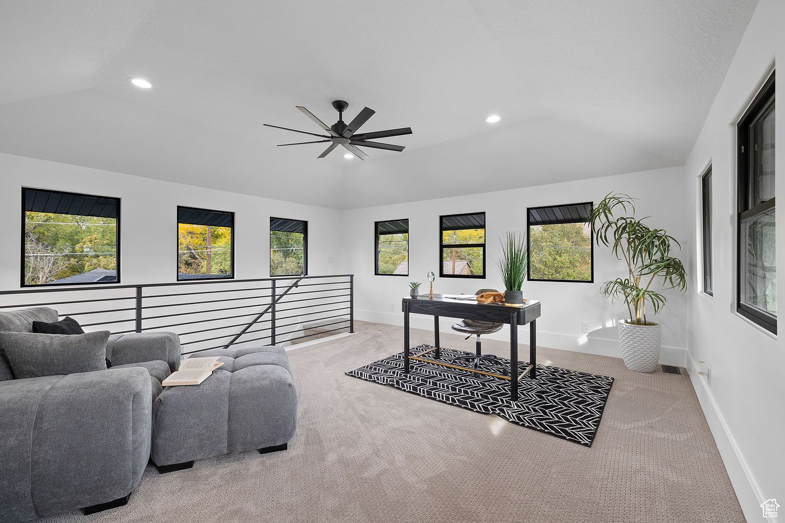This is an interior shot of a modern office space featuring a black desk with gold accents, a patterned rug, and a gray sofa with ottoman. The room is well-lit with natural light from multiple windows, complemented by recessed lighting and a ceiling fan. A large potted plant adds a touch of nature to the space, creating a bright and inviting atmosphere.