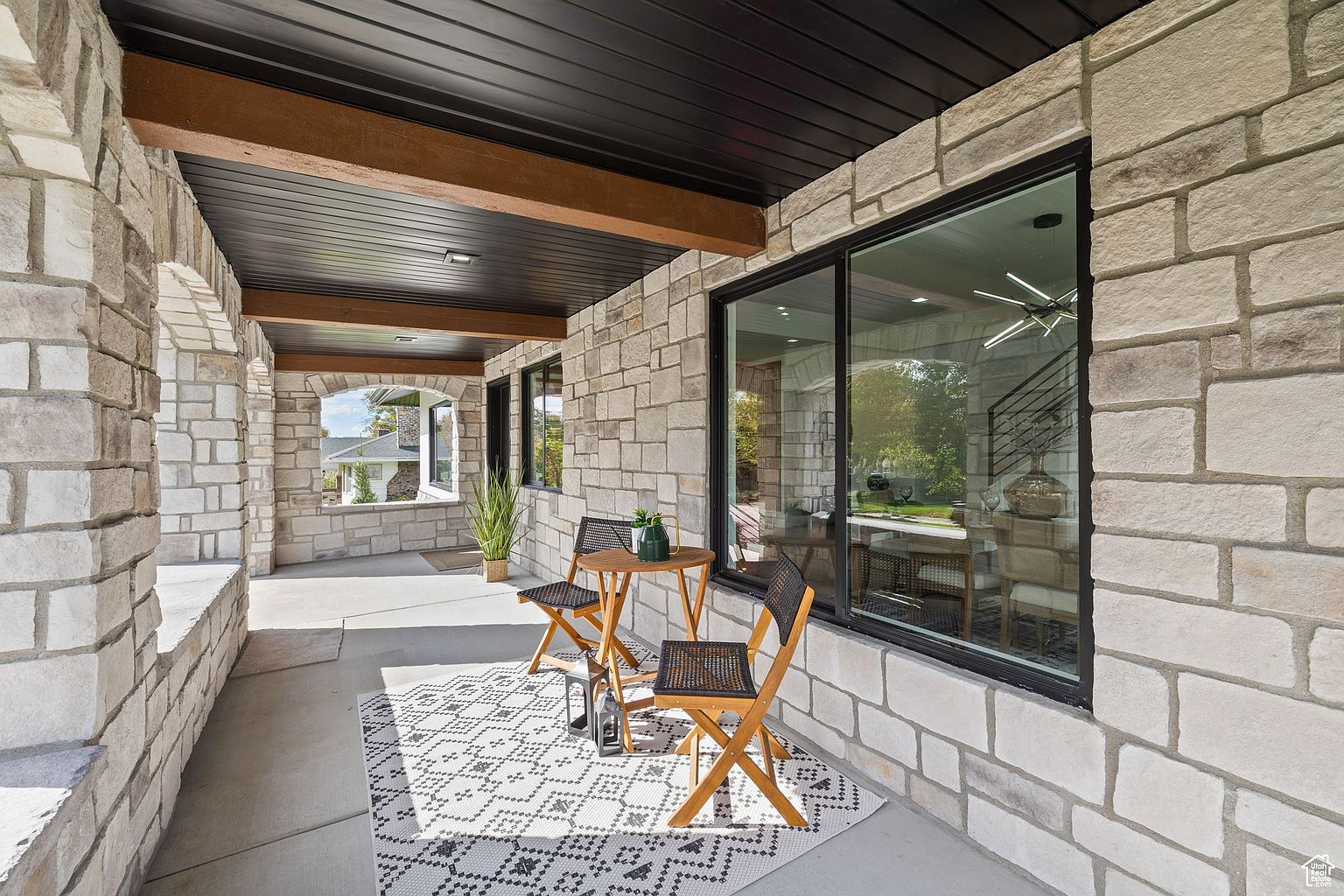 This image showcases a charming outdoor patio area, featuring a stone facade, a black ceiling with wooden beams, and a stylish seating arrangement with a small round table and two chairs on a patterned rug. The space is well-lit and inviting, offering a perfect spot for relaxation or entertaining.