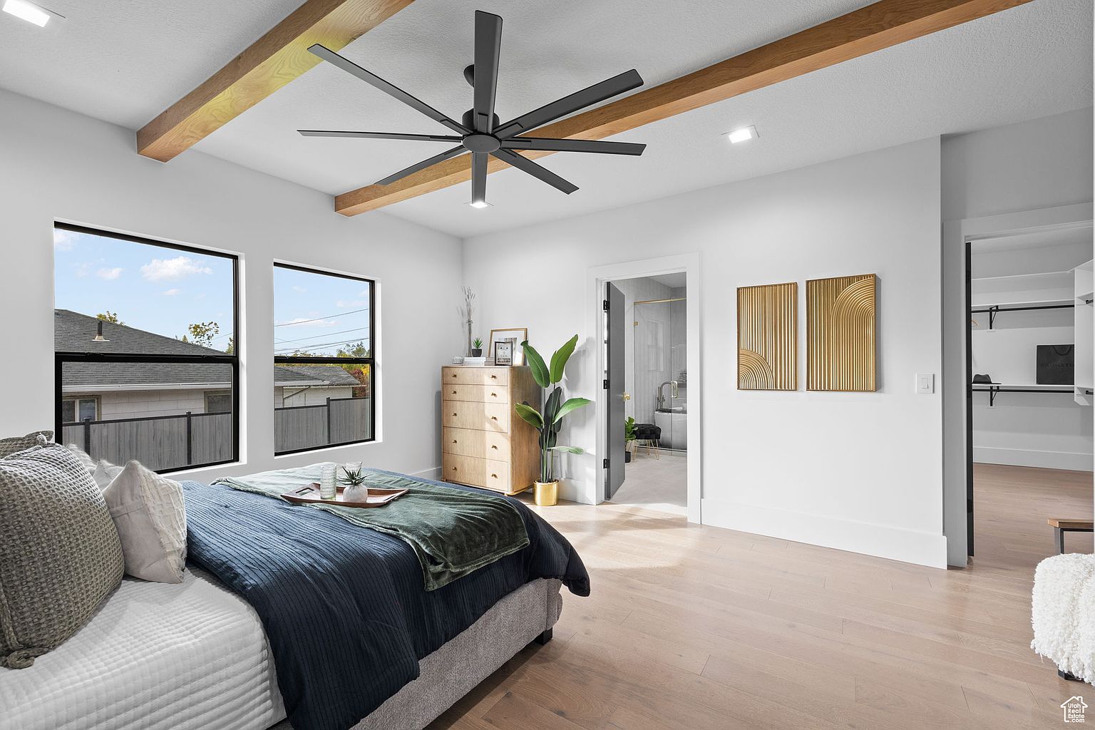 This is a bright and airy primary bedroom featuring a modern aesthetic. The room includes a bed with a blue duvet and decorative pillows, two windows providing natural light, and exposed wooden beams on the ceiling. A ceiling fan, a dresser, and artwork add to the room's appeal, while an open doorway leads to a walk-in closet and another to a bathroom.