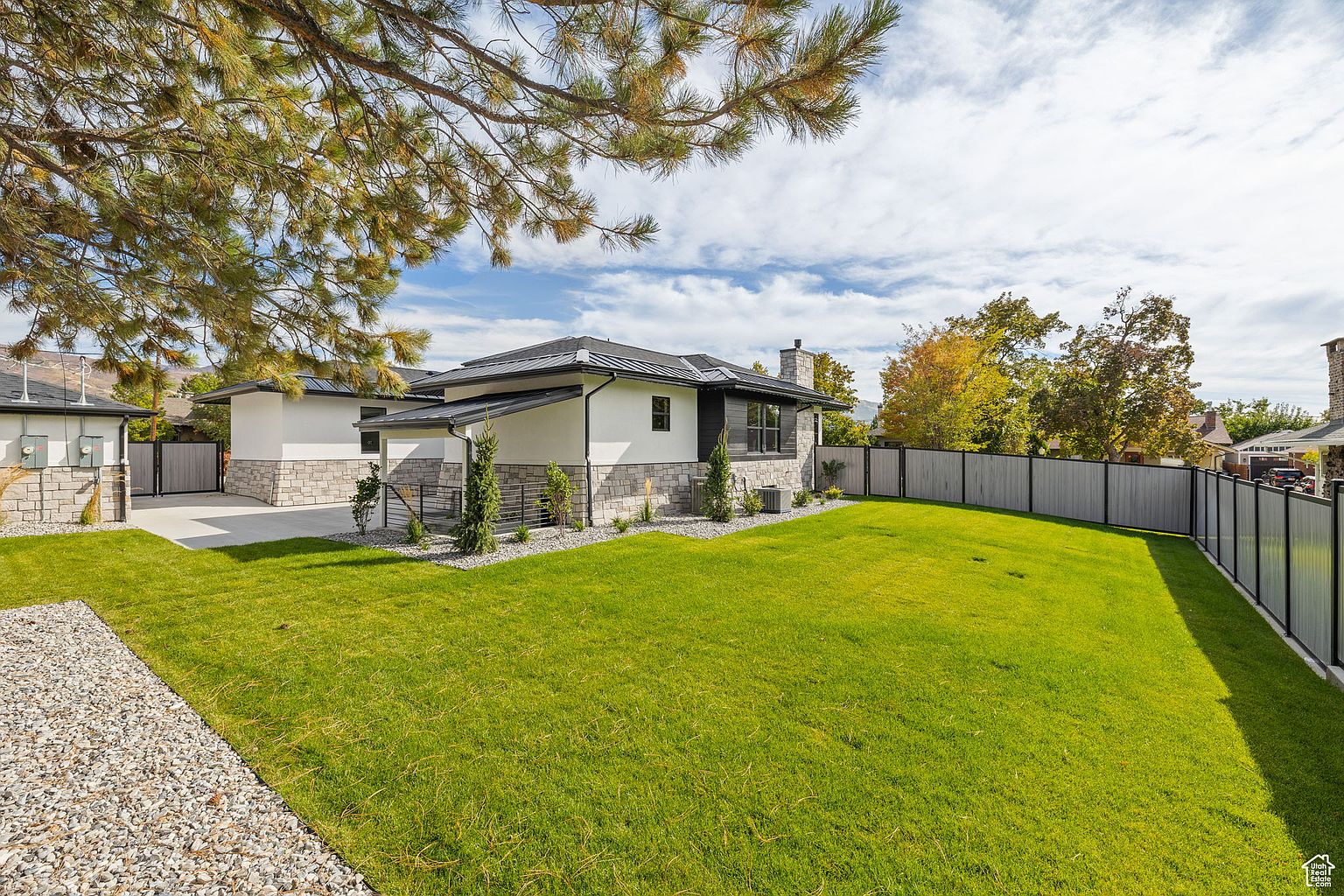 This image showcases the side of a modern home with a well-manicured lawn. The house features a combination of stone and white siding, a dark roof, and neatly trimmed landscaping. A gray fence runs along the property line, adding privacy and defining the outdoor space.