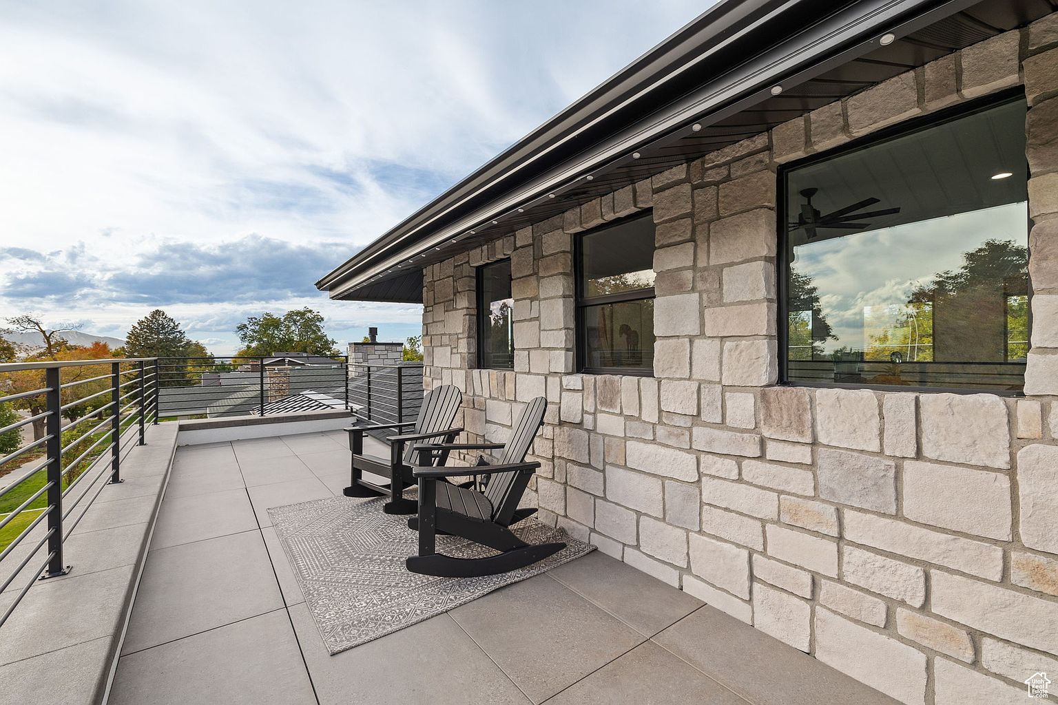 This image showcases a modern balcony featuring two black Adirondack chairs on a gray patterned rug, set against a stone-clad exterior wall with black-framed windows. The balcony has a sleek, horizontal railing offering a view of the landscape, and the overall design exudes a contemporary and sophisticated aesthetic. The perspective is from a medium distance, capturing the balcony's layout and its connection to the surrounding environment.